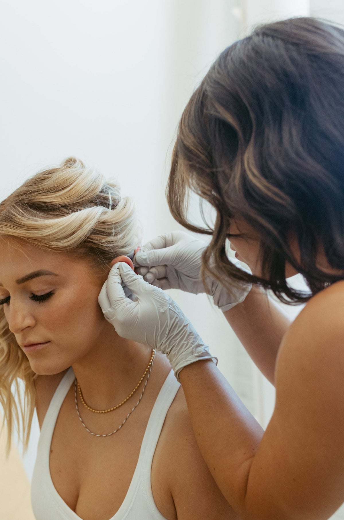 A woman with blonde hair sits while another woman wearing white gloves adjusts an ear cuff on her ear. The seated woman wears a white ribbed tank top and light blue jeans in a bright indoor setting.